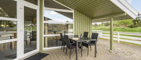 An outdoor patio with a table and chairs, a covered area with a roof, and a white fence in the background.