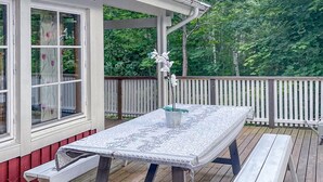 Um pátio externo com mesa e bancos, rodeado por um deck de madeira e um corrimão branco.