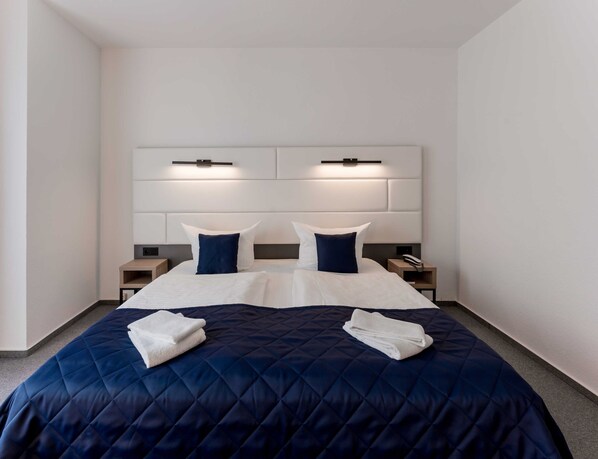 A modern hotel room with a large bed, two bedside tables, a headboard with integrated lighting, and a neutral color scheme.