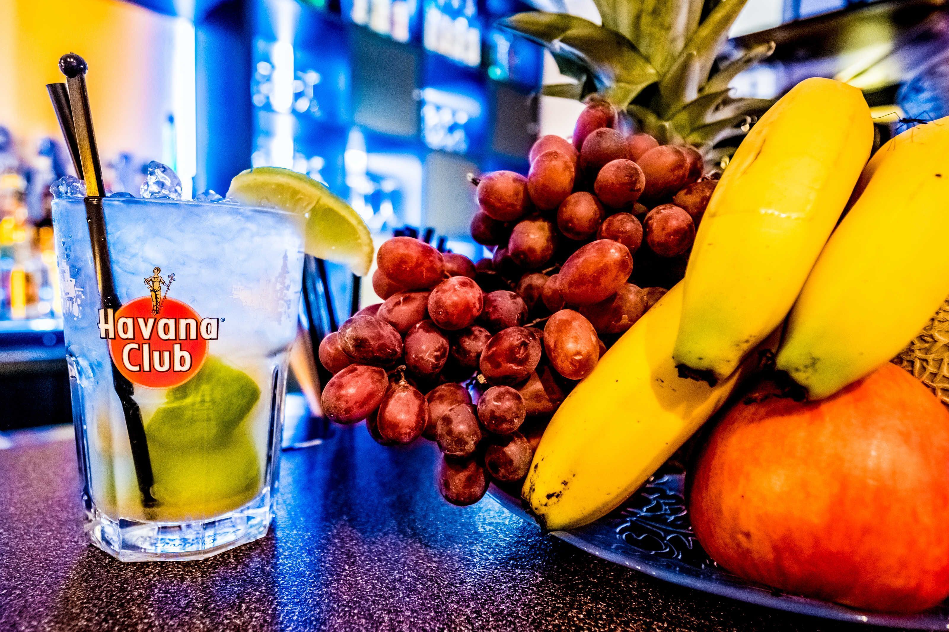 A glass of Havana Club rum with a lime wedge and a straw, accompanied by a plate of bananas, grapes, and a pineapple.