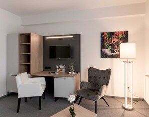 Et moderne hotelværelse med et skrivebord, to stole, et fjernsyn, et billede og en gulvlampe.