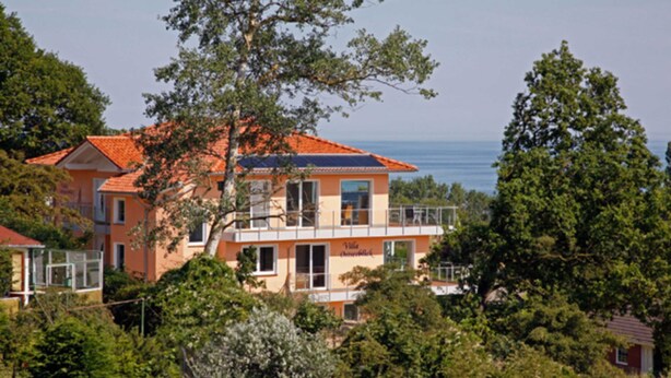 Un bâtiment à deux étages, doté d’un toit recouvert de tuiles rouges, de grandes fenêtres et d’un balcon. Il est entouré de verdure et offre une vue sur une étendue d’eau.