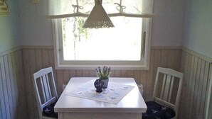 A small dining area with a white table, two white chairs, a lace chandelier, and a window with a view of trees.