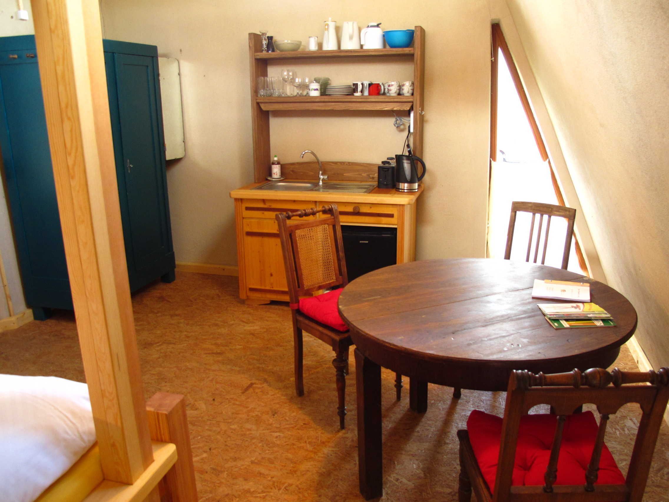 A small, cozy room with a wooden dining table, chairs, a kitchenette with shelves above the sink, and a window providing natural light.