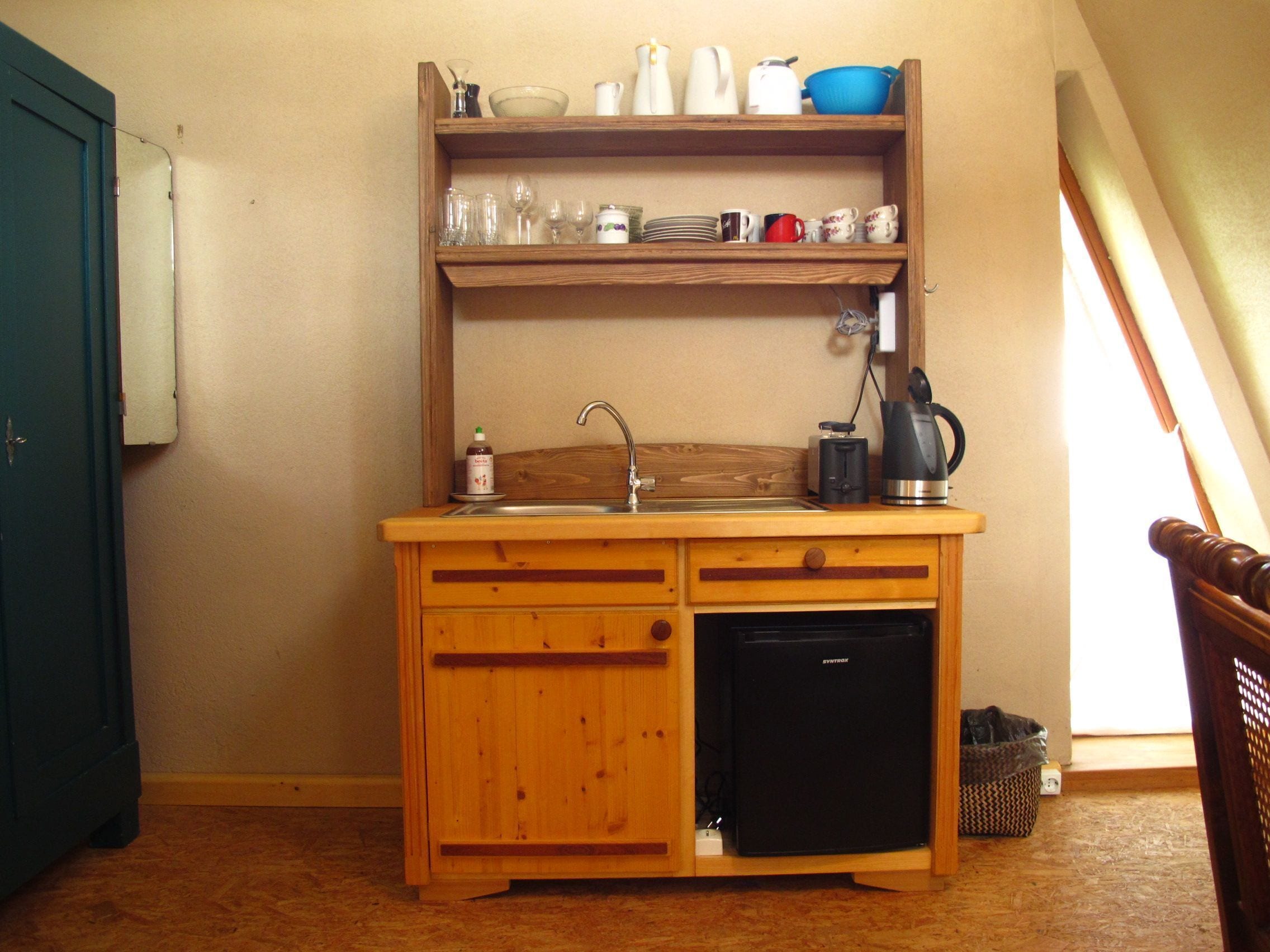 A compact kitchenette with a sink, a small refrigerator, and a shelf above holding various kitchenware.