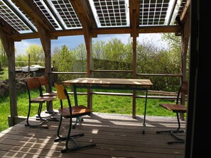 Ein überdachter Außenbereich mit Tisch und Stühlen, Solaranlagen auf dem Dach und Blick auf Bäume und Rasen.