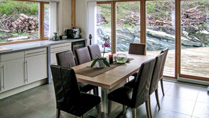 Een moderne keuken met grote ramen, een eettafel met stoelen en uitzicht op een bebost gebied.