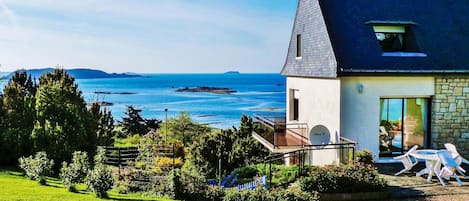 A house with a balcony overlooking a sea view, a garden with various plants, and outdoor seating.