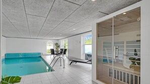 Een modern binnenzwembad met een helder uitzicht op het zwembad, een zithoek met stoelen en een glazen scheiding met een houten bankje.