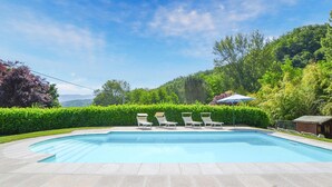 Una piscina rectangular con una terraza de baldosas, rodeada por un seto bajo y una fila de sillas reclinables blancas.