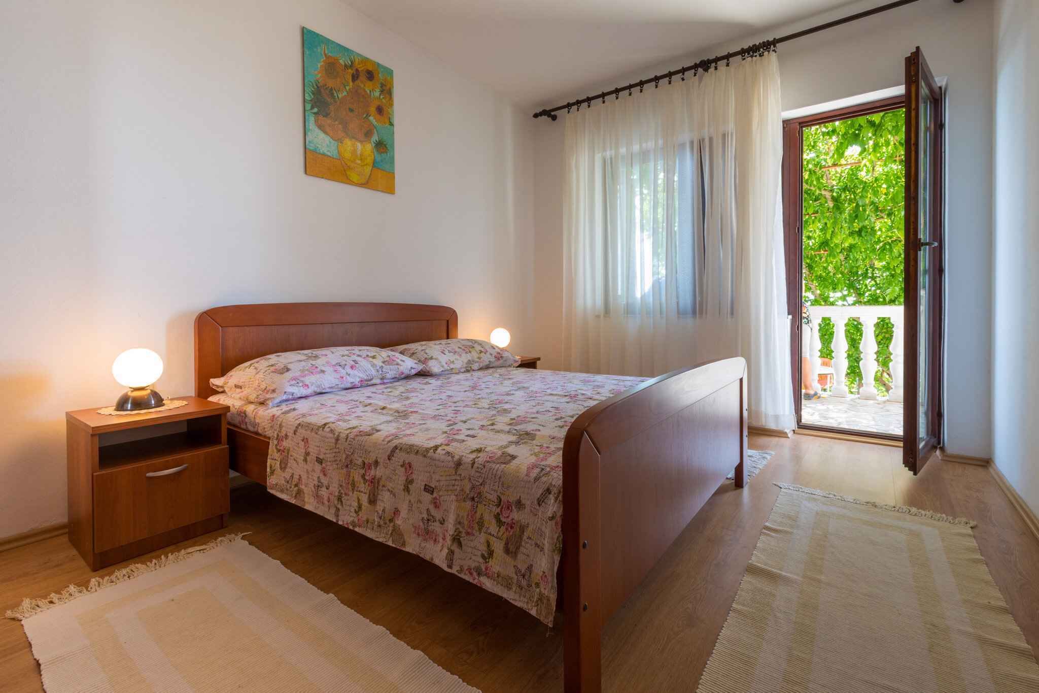 A bedroom with a wooden bed, bedside tables, a nightstand, a window with curtains, and a view of greenery outside.