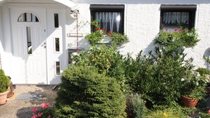 A white building with a white door, two windows with curtains, and a small garden area with potted plants and flowers. - Apartment Bergblüte - FeWo (Bad Harzburg)