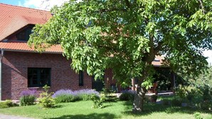 A brick building with a red roof, surrounded by a well-maintained garden with various plants and a large tree. - Kräuter-Landhaus, Germany (Bad Schmiedeberg)