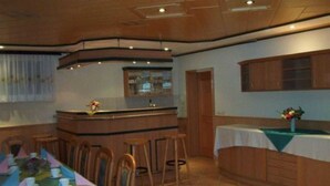 A dining area with a long table set for a meal, wooden chairs, a bar area with stools, and a kitchen in the background. - Kräuter-Landhaus, Germany (Bad Schmiedeberg)