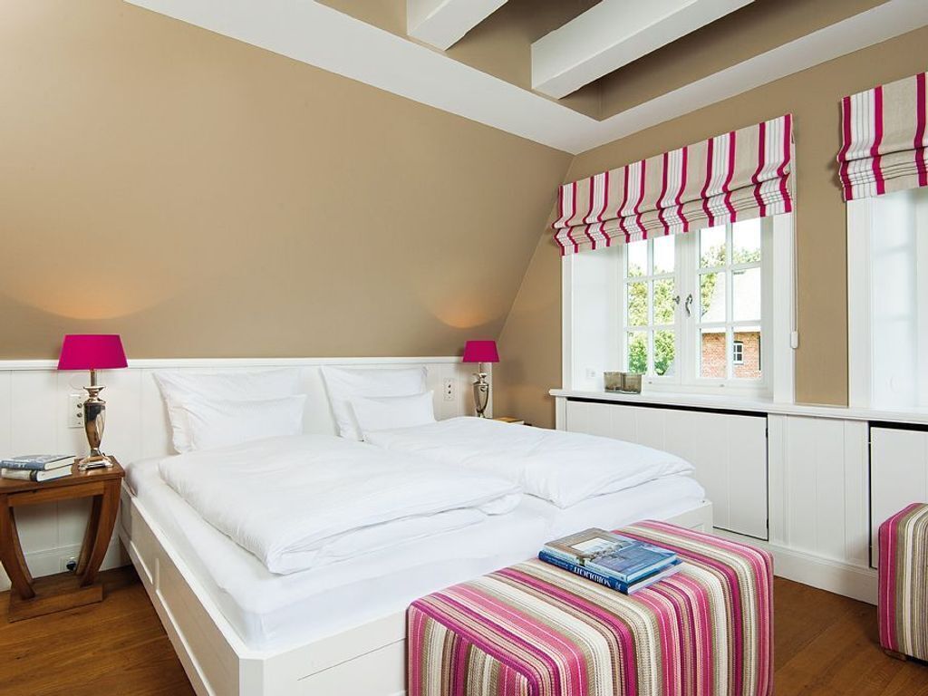A bedroom with a large bed, two bedside lamps, a window with striped valance, and a small bedside table with books.