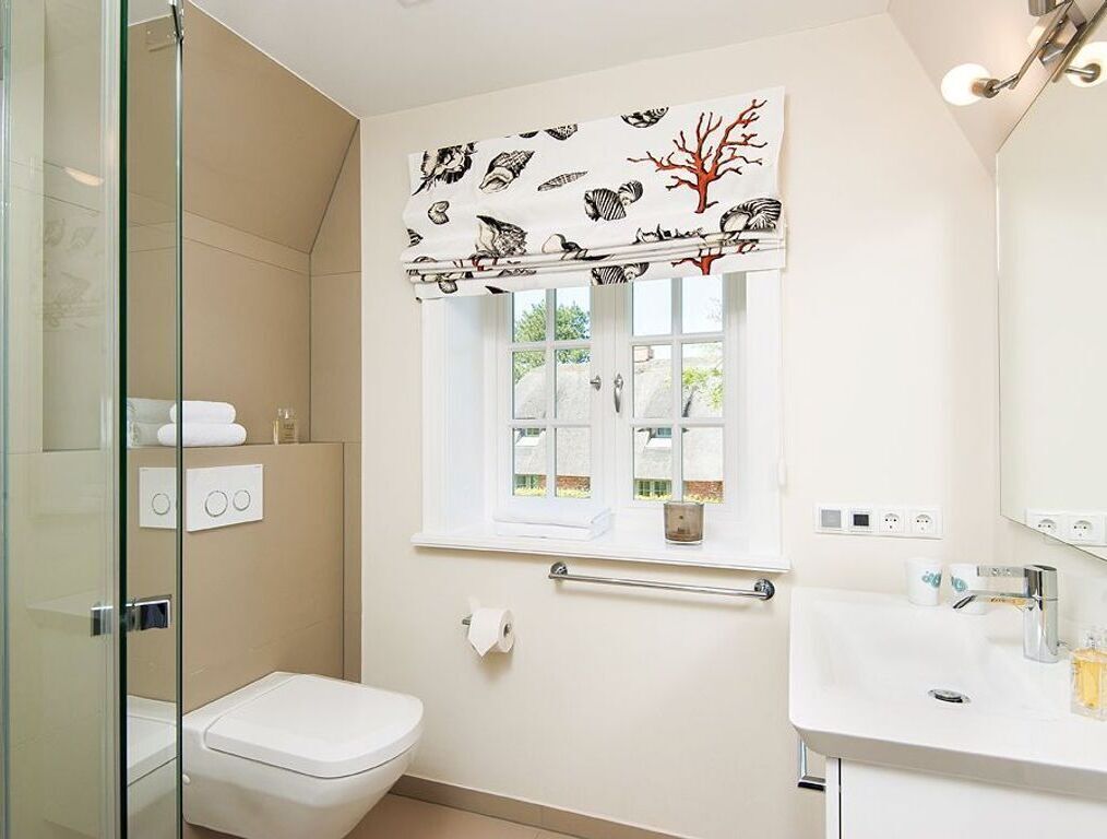 A bathroom with a toilet, a sink, a mirror, a window with a curtain, and a shower area.