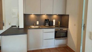 A compact kitchen with white cabinets, a black countertop, a coffee maker, a kettle, and an oven. - (171) 3- room apartment - apartment house Strandstr. 16 (Kühlungsborn)