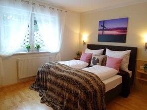 A bedroom with a bed, a window with curtains, a radiator, a wall-mounted light, and a painting.