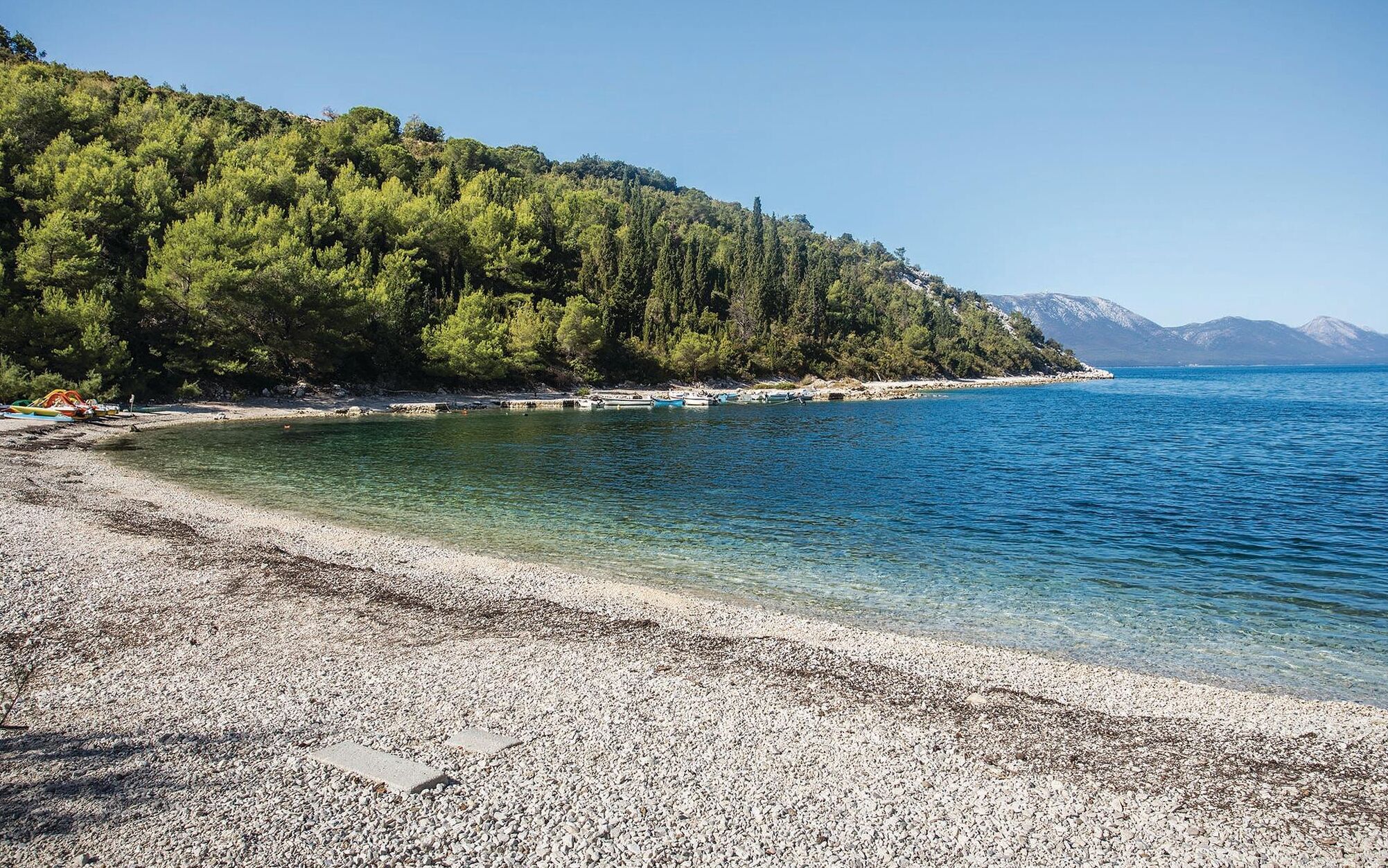 A pebble beach with clear water, boats, and a forested hill.