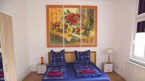 Une chambre à coucher avec un lit double, deux tables de chevet, une commode, une fenêtre avec des stores et un tableau au mur.