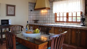 Una cocina con una mesa de comedor, sillas, microondas, un lavamanos y un ventanal con cortinas.