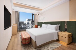 Una habitación de hotel moderna con una cama grande, una mesita de noche, un banco y vistas a la ciudad desde un balcón.