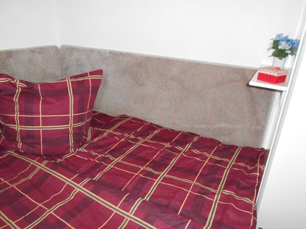Una cama en esquina con colcha a cuadros rojos y dorados, un almohadón y una pequeña estantería con una caja roja y una planta.