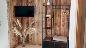 Ein Zimmer mit einer Holzwand, einem flachbildfernseher über einer schwarzen Metallregaleinheit und einer Vase mit Trockenpflanzen.