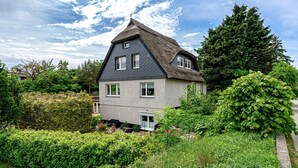 Ein Haus mit einem steilen Dach, einer kleinen Veranda und einem Garten mit verschiedenen Pflanzen und Blumen.