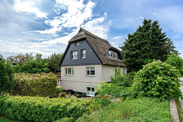 Une maison avec un toit à forte pente, un petit porche et un jardin où poussent différentes plantes et fleurs.