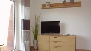 Huoneessa on televisio, joka on sijoitettu puiseen kaappiin, ruukussa oleva kasvi ja hylly, jossa on koriste-esineitä.