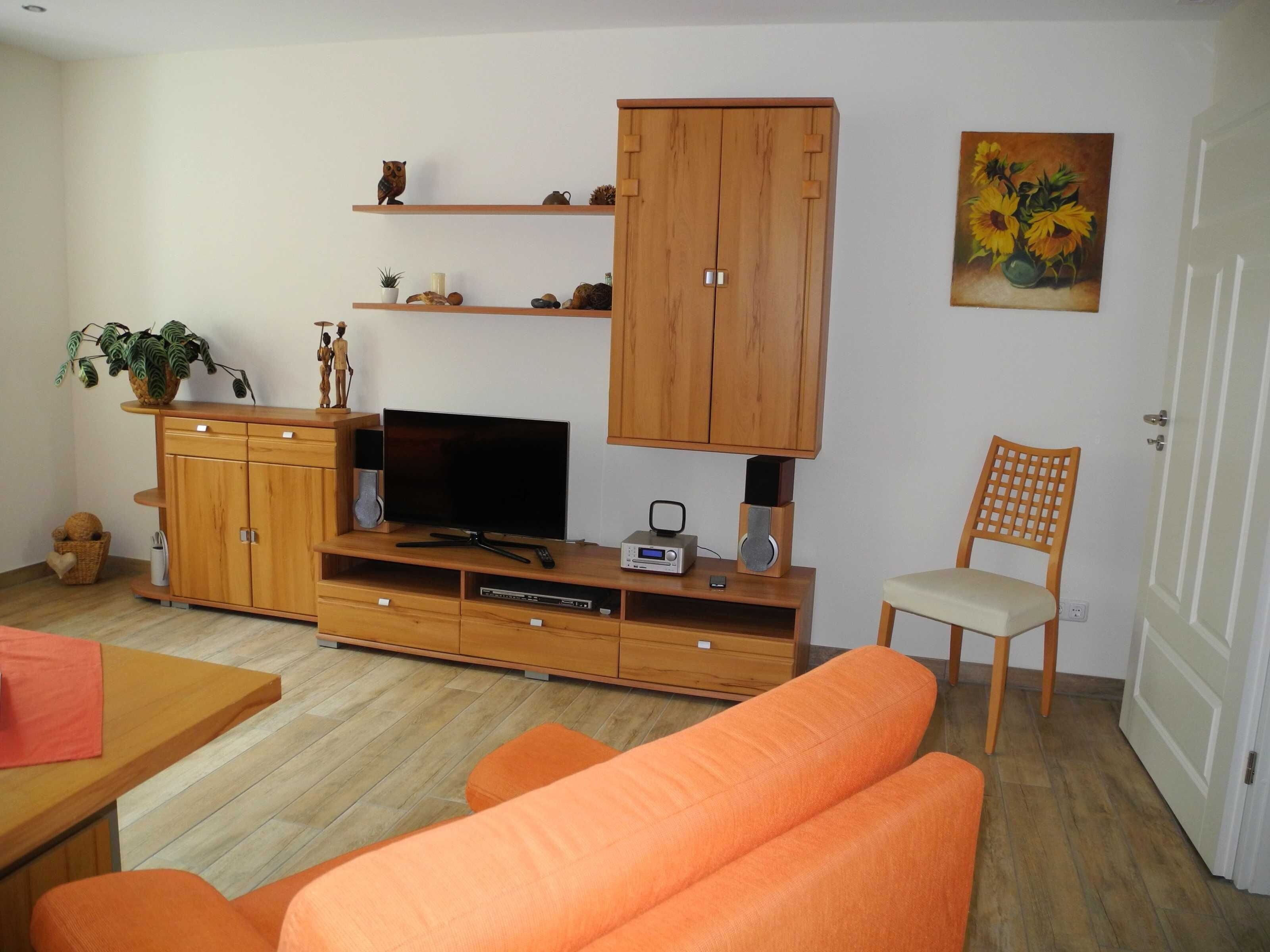 Un salon avec un téléviseur, un meuble de rangement en bois, un canapé orange et un tableau représentant des tournesols.