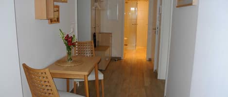 A hallway with a wooden dining table and chairs, a vase with flowers, a wooden cabinet, and a bathroom visible in the background.