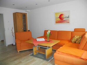 Ein Wohnzimmer mit einem orangefarbenen Sofa, einem Holzschrank, einem kleinen Tisch mit Blumen und einem Bild an der Wand.