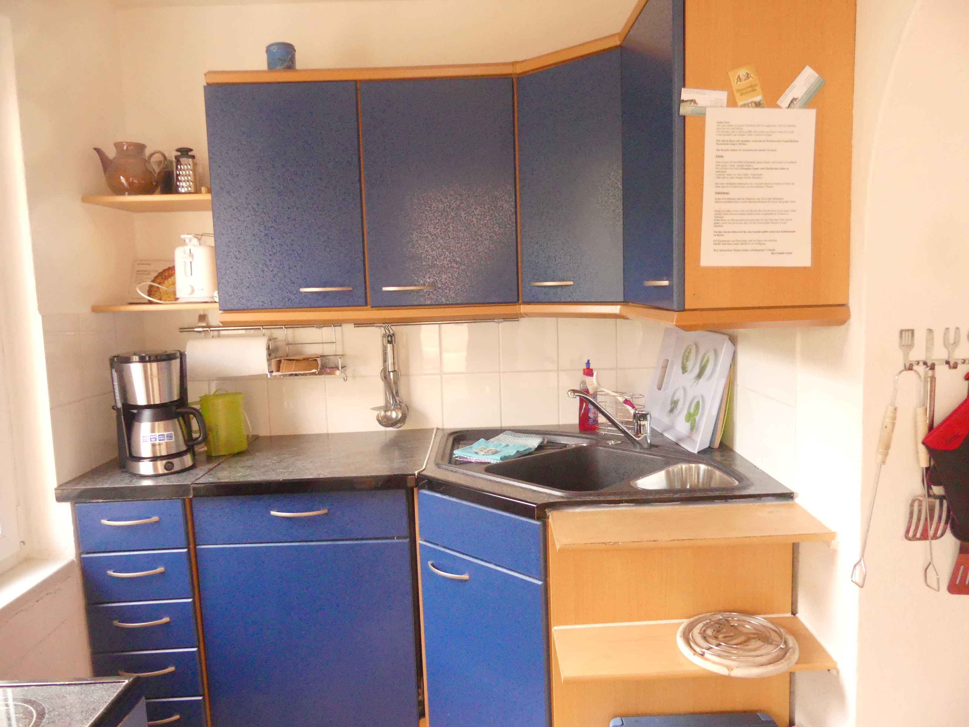Een compacte keuken met blauwe kastjes, een koffiezetapparaat, een spoelbak en een klein schap met keukengerei.
