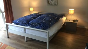 A bedroom with a bed, two bedside lamps, a nightstand, a painting on the wall, and a window with curtains. - Schiel apartment rental - Apartment (Zempin)