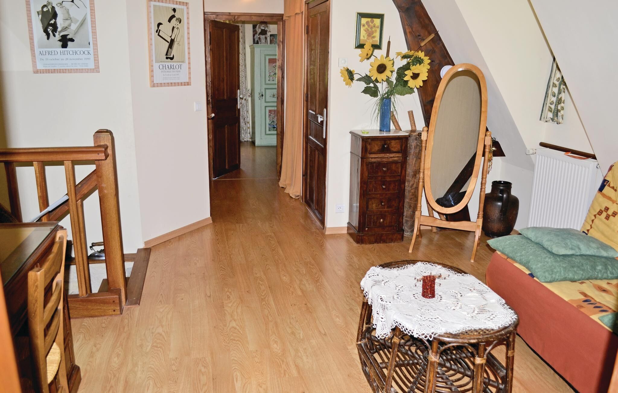 Ein gemütliches Zimmer mit Holzboden, einem kleinen runden Tisch mit weißer Spitzenunterlage, einem Spiegel, einer Vase mit Sonnenblumen und einer Couch mit gemustertem Kissen.