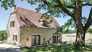 Una casa in pietra con tetto in tegole, un grande albero di fronte e un patio con mobili da esterno.