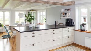 Et moderne køkken med hvide skabe, en sort bordplade og en central ø med skuffer. Der er planter på øen, en kaffemaskine og en hylde med dekorative genstande.