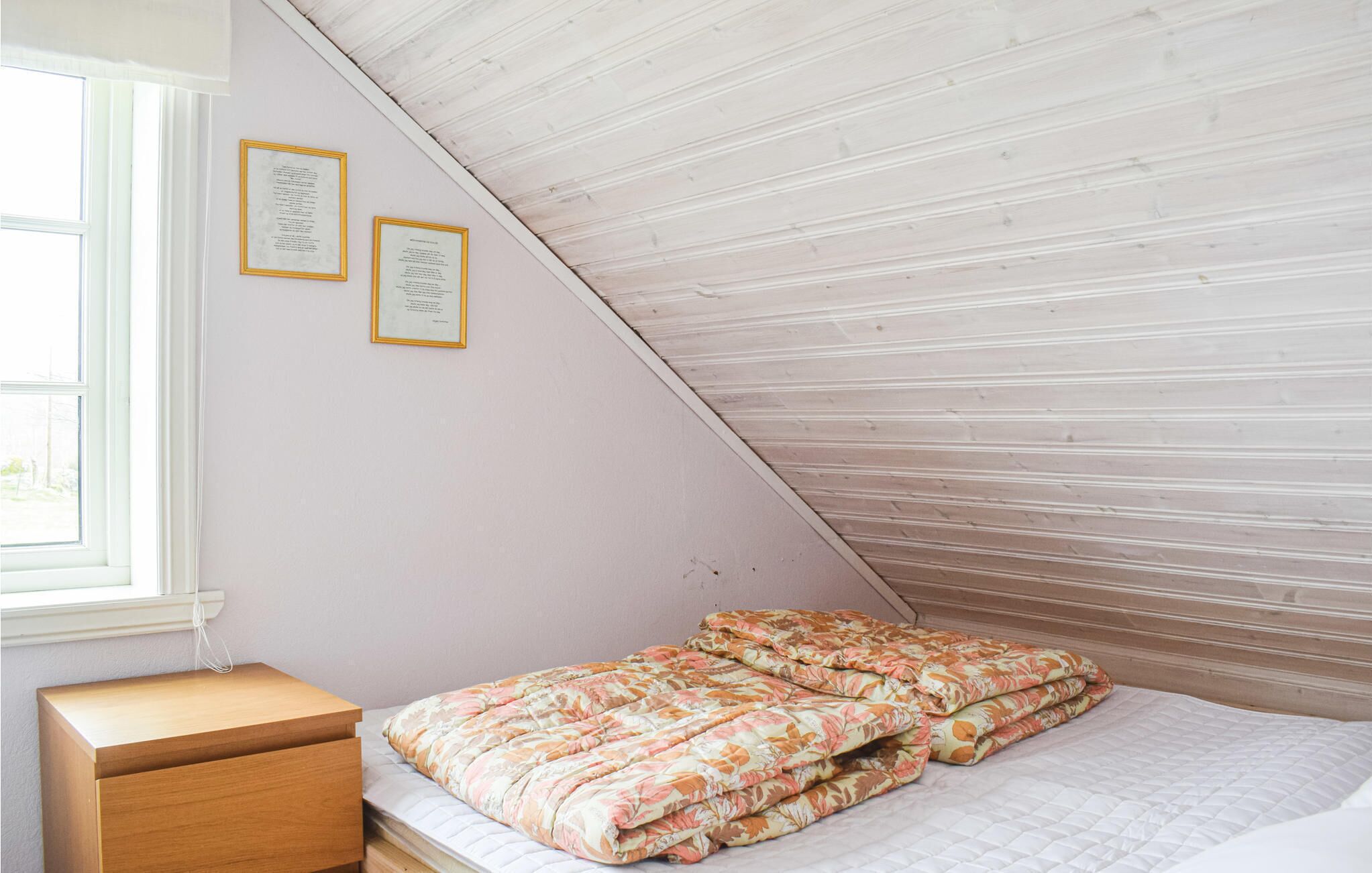 Un dormitorio con techo inclinado, una mesita de noche de madera, una cama con edredón floral y dos diplomas enmarcados en la pared.