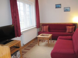 Ein Wohnzimmer mit Fernseher auf einem Holzständer, einem roten Sofa mit zwei Kissen, einem kleinen Holztisch und einer Heizung.
