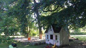 A small wooden playhouse with a heart-shaped window, surrounded by trees and a green lawn. - Vacation apartment Möwe (ground floor) - Windland Ranch - Equestrian farm with vacation apartments (House (Altenkirchen)