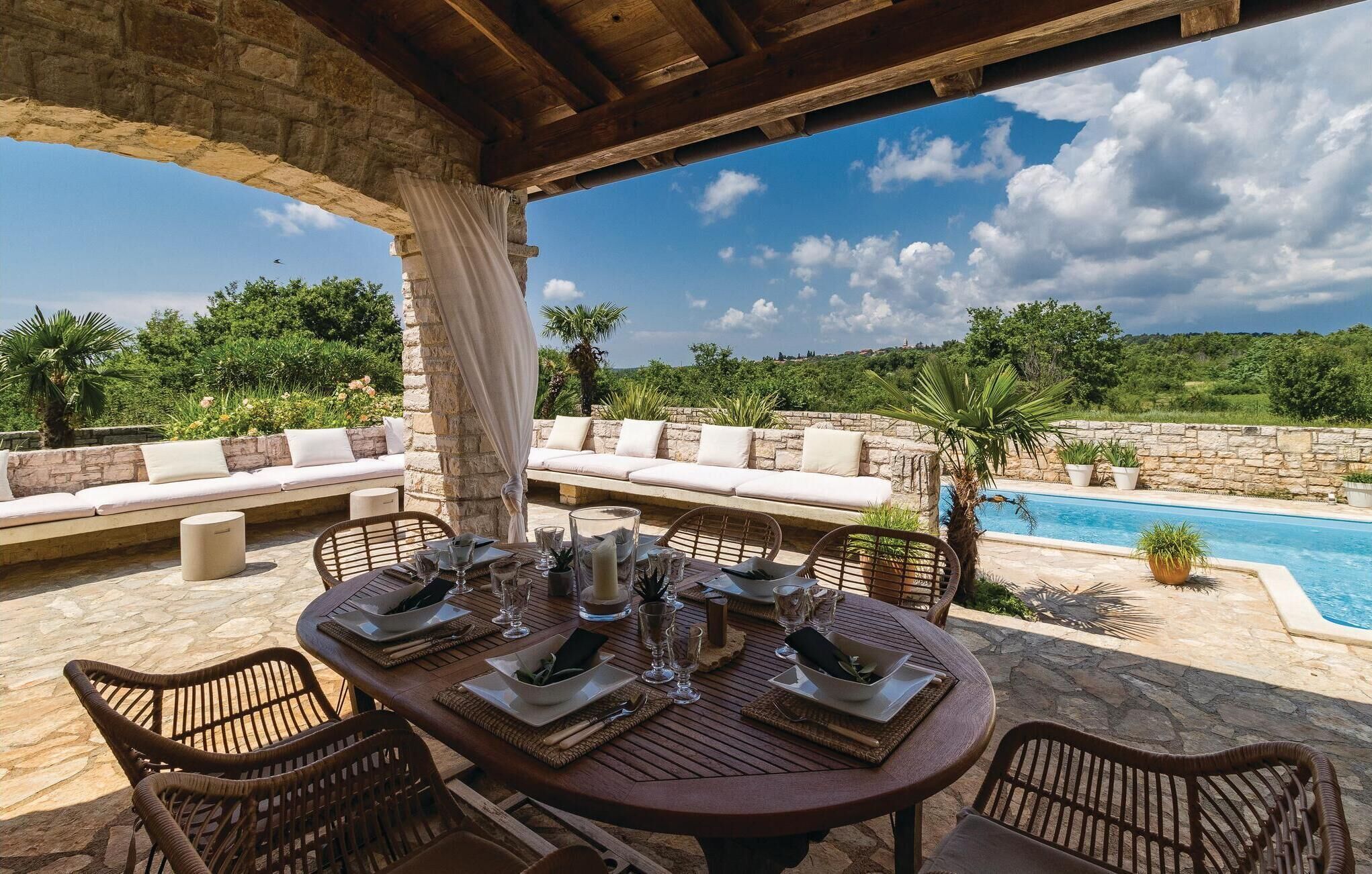 A covered outdoor dining area with a stone table and wicker chairs, overlooking a pool and lush greenery.