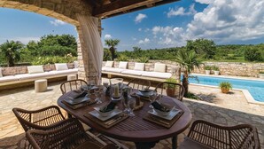 A covered outdoor dining area with a stone table and wicker chairs, overlooking a pool and lush greenery.