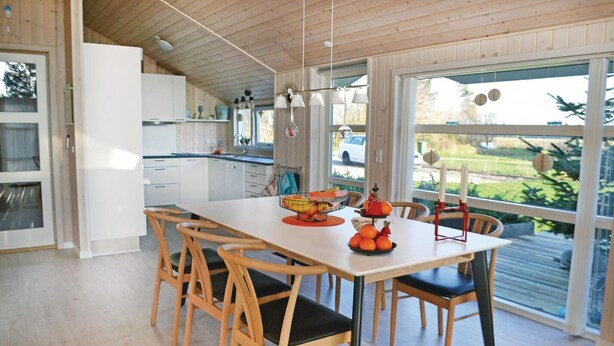 Eine moderne Küche mit weißen Schränken, einem Holztisch und Stühlen. Auf dem Tisch steht eine Obstschale und ein Fenster bietet Blick ins Freie.