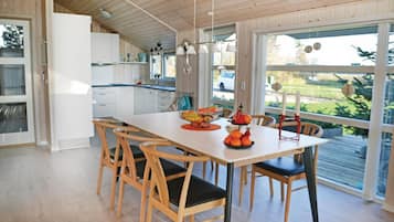 Une cuisine moderne avec des meubles blancs, une table à manger en bois et des chaises. On peut voir un panier de fruits sur la table, et une fenêtre donnant sur l’extérieur.