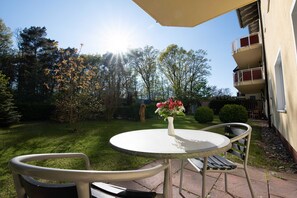 Une terrasse avec une table ronde et deux chaises, un vase rempli de fleurs, et une vue sur un jardin et un bâtiment en arrière-plan.