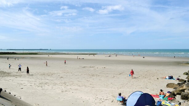 A wide sandy beach with people walking and playing, a tent set up, and a calm sea in the background.