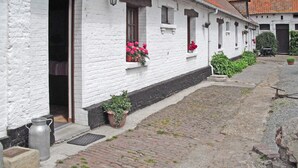 A white building with a brick foundation, a wooden door, and windows with flower boxes. There are potted plants along the pathway and a cobblestone street.