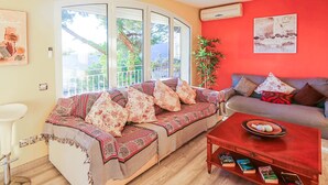A cozy living room with a sofa adorned with patterned cushions, a red coffee table with books, and a view of a balcony with greenery. - Gorgeous home in Santa Susanna (Santa Susanna)